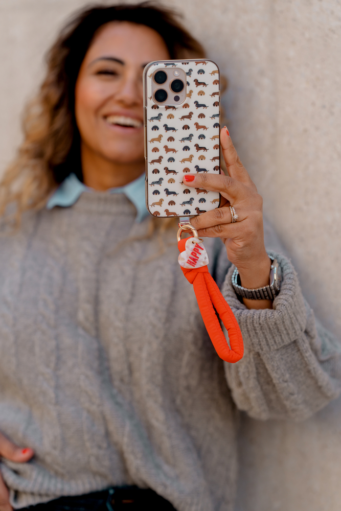 Smartphone in Hülle mit Dackelmotiven und orangefarbener Handschlaufe mit Anhänger in einer Hand gehalten.
