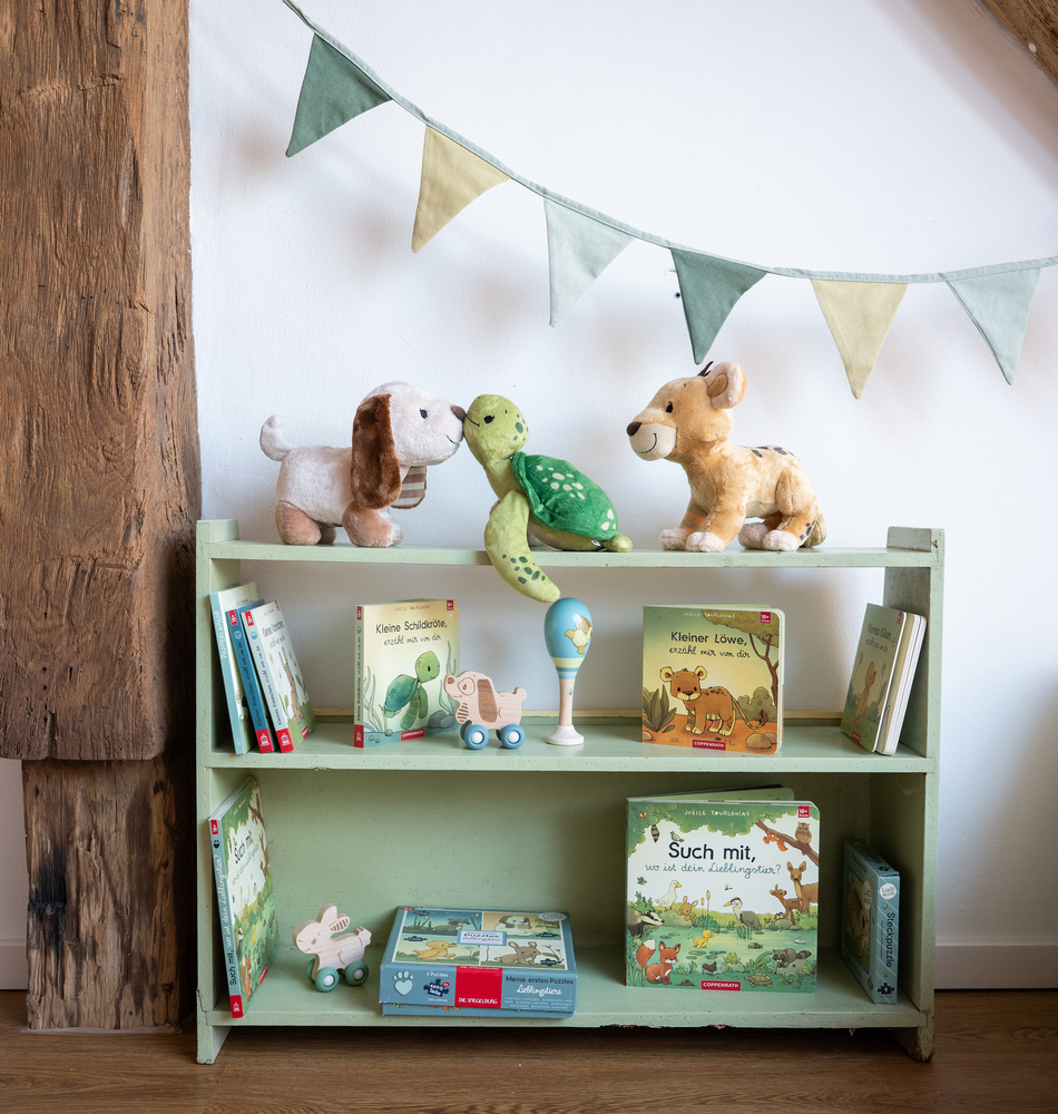 Grünes Regal mit Kinderbüchern, Stofftieren und Holzspielzeug, dahinter Wimpelkette in Grün- und Beigetönen.