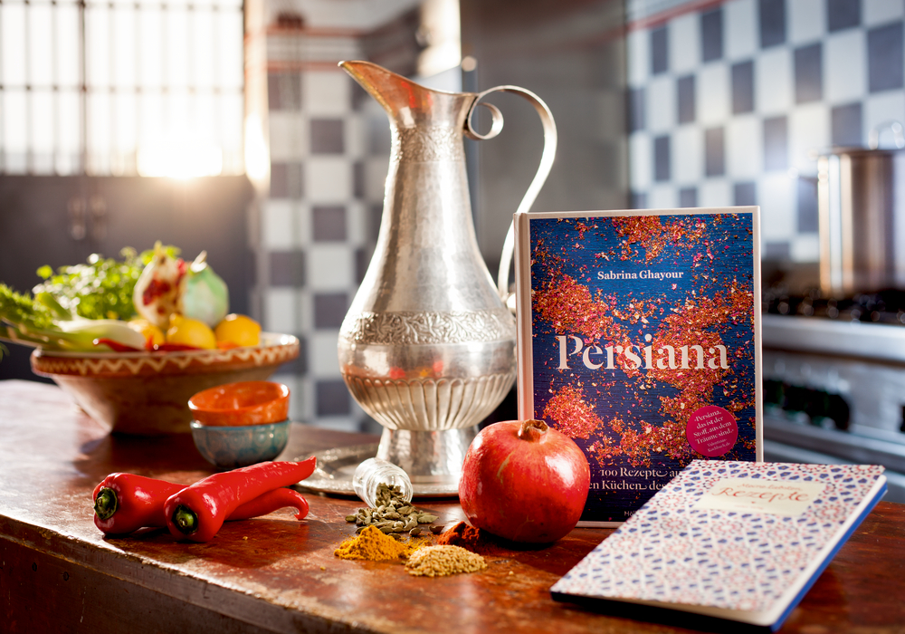 Ein Kochbuch, Gewürze, Granatäpfel und rote Paprika auf einer rustikalen Theke neben einem silbernen Krug.