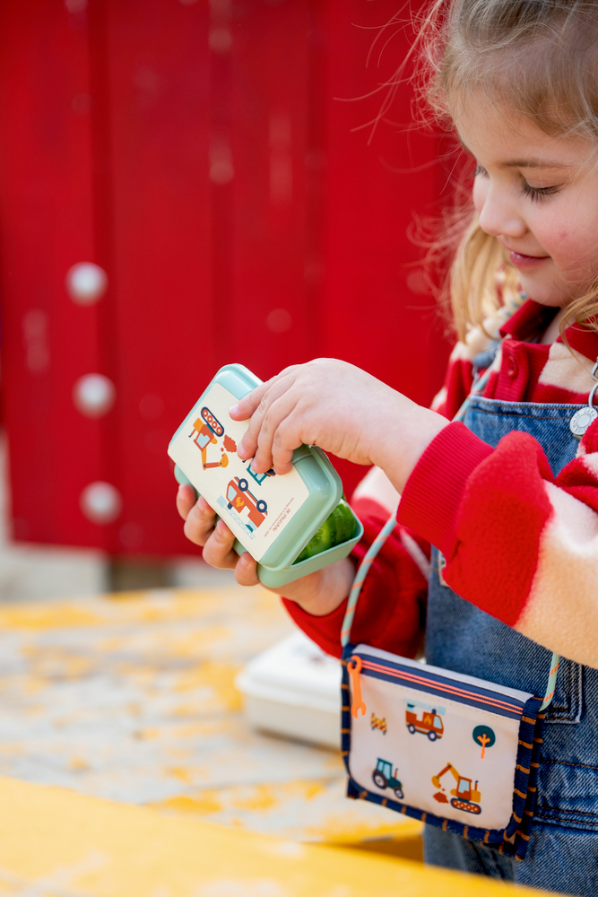 Kind hält türkisene Mini-Snackbox mit Fahrzeugmotiven, trägt rot gestreiften Pullover, Jeanslatzhose und kleine Umhängetasche mit Baustellenmotiven.