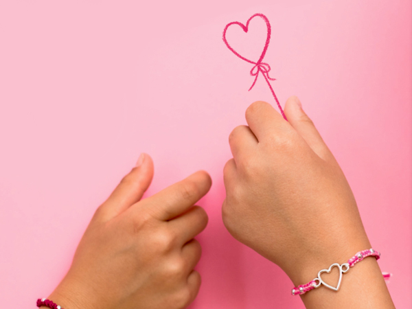 zwei Kinderhände nebeneinander, die linke trägt ein kleines Rosa Armband mit Herzanhänger und hält eine Herzförmige pinke Schnur in der Hand