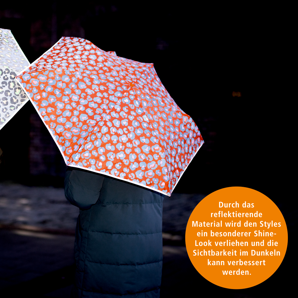 Person mit orange und reflektierend gemustertem Regenschirm in dunkler Umgebung, daneben teilweise ein zweiter Schirm sichtbar.