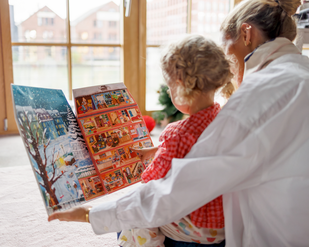 Erwachsene und Kind betrachten gemeinsam einen geöffneten Adventskalender mit winterlichem Motiv und Innenansicht eines Hauses.