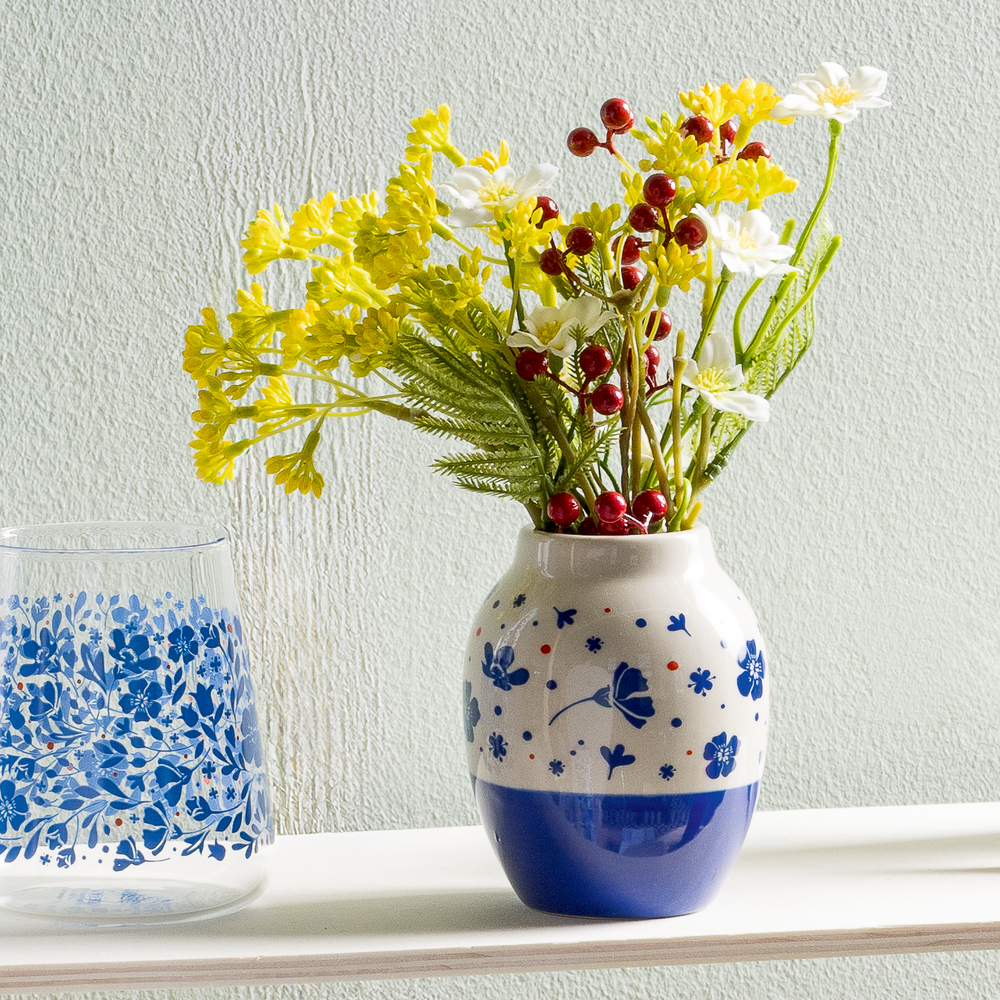 Beige Keramikvase mit blauem Blumendekor, darin gelbe und weiße Blumen mit roten Beeren, daneben ein Glas mit blauem Muster.