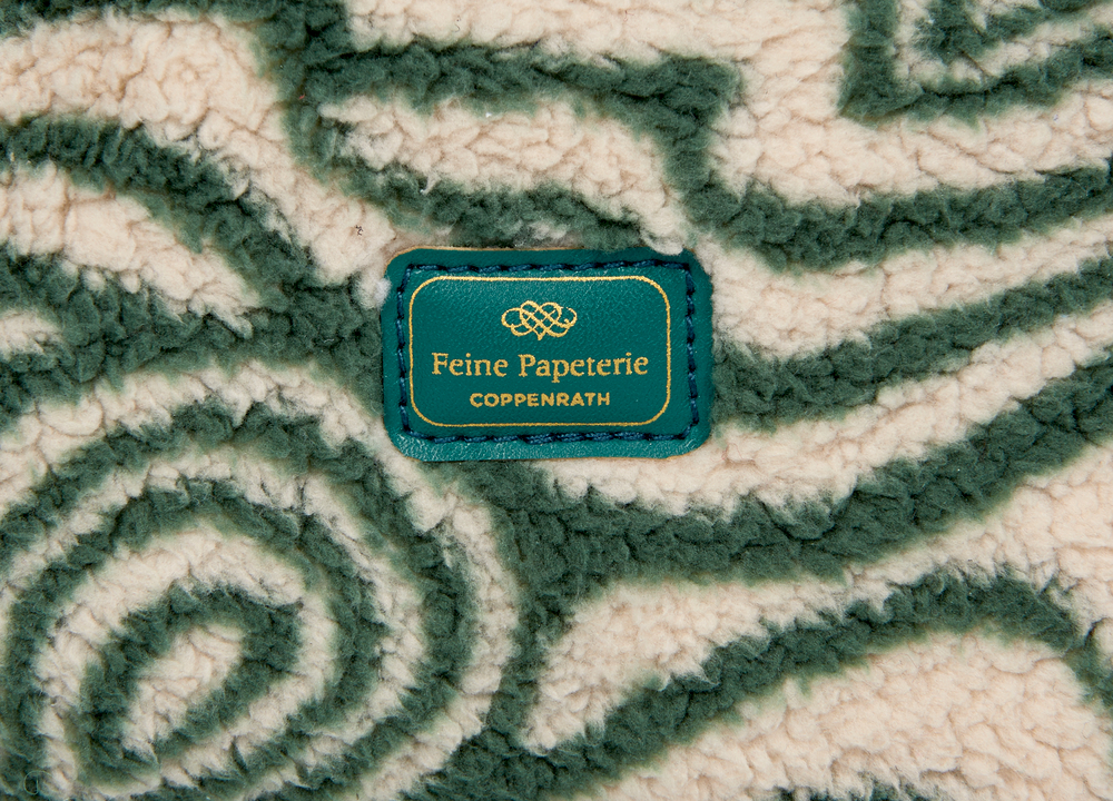 Grüne Stoffoberfläche mit cremefarbenem Muster und grünem Etikett mit goldener Aufschrift „Feine Papeterie Coppenrath“.