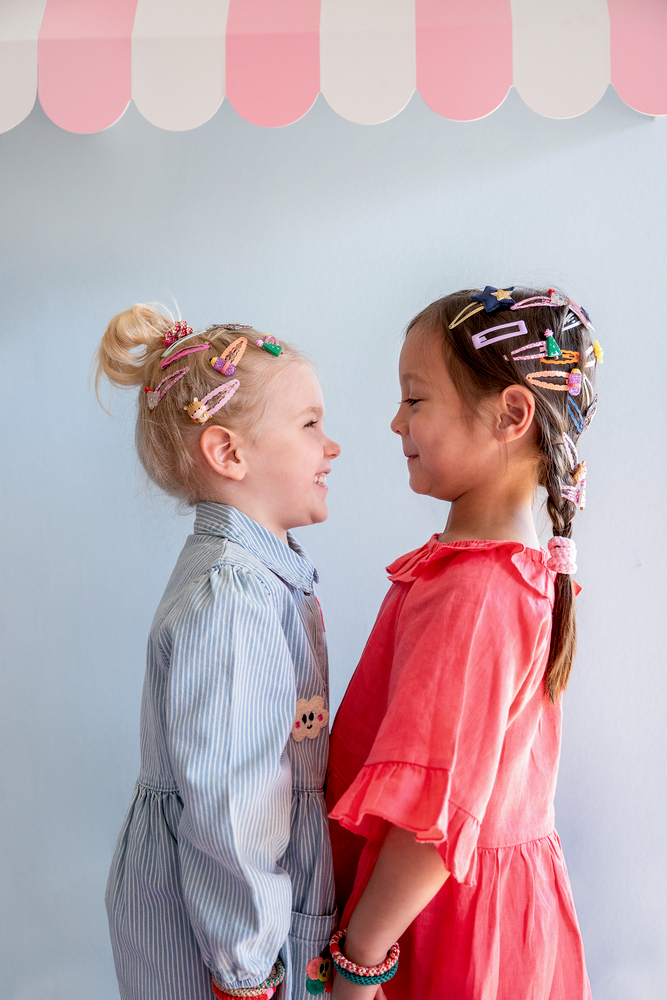 Zwei Kinder stehen sich gegenüber, tragen bunte Haarspangen und Armbänder sowie Kleider in Blau und Rosa.