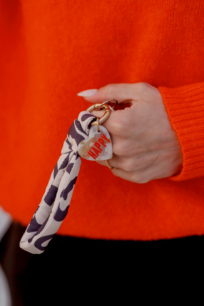 Hand mit orangefarbenem Ärmel hält Schlüsselanhänger mit gemustertem Stoffband und herzförmigem Anhänger mit rotem Schriftzug „HAPPY“.