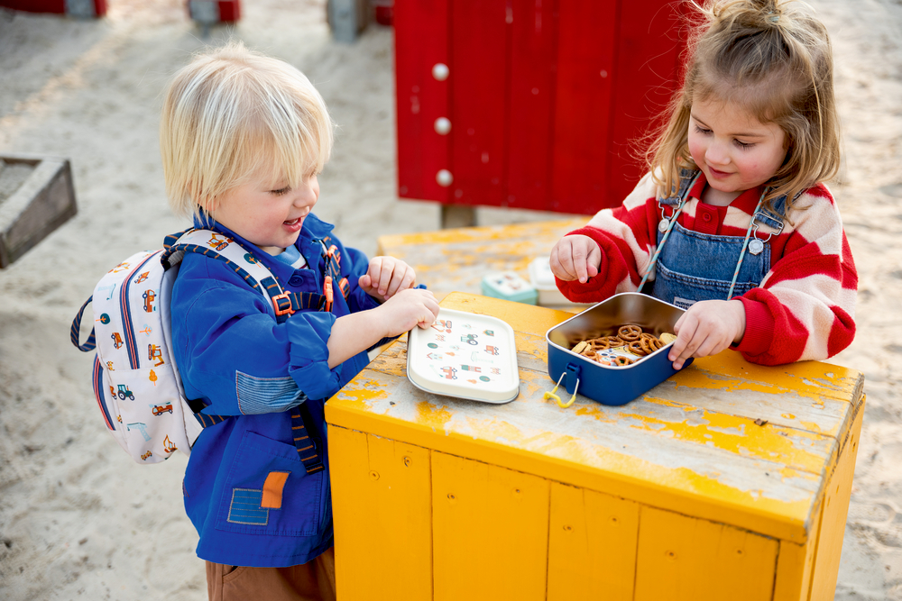 Zwei Kinder essen an einem gelben Holztisch aus der Brotdose mit Salzbrezeln im Freien.